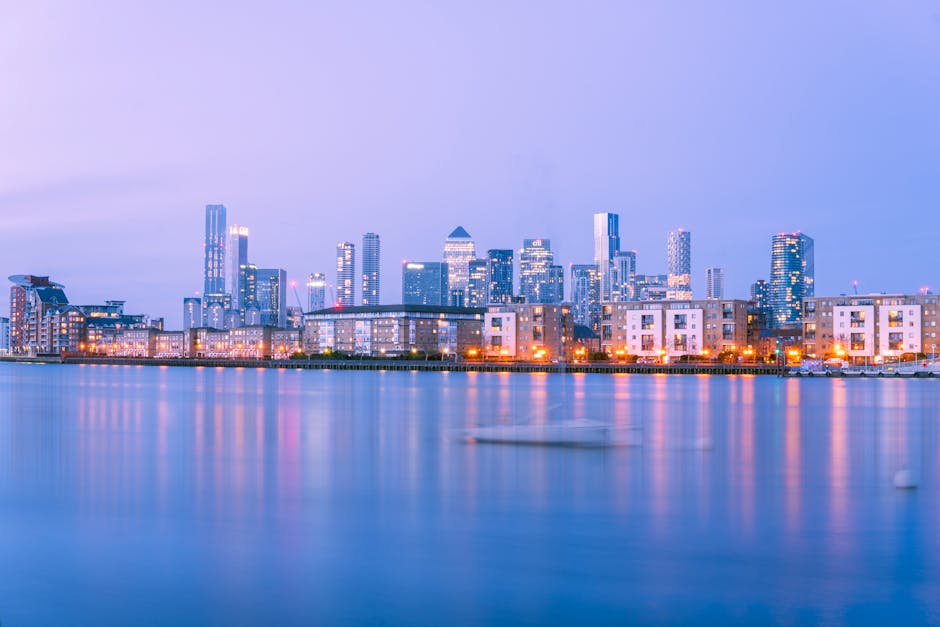 A view of tall modern office buildings situated along a river in an urban cityscape, with a clear blue sky above. The buildings feature glass facades reflecting the sky and surrounding structures, wit
