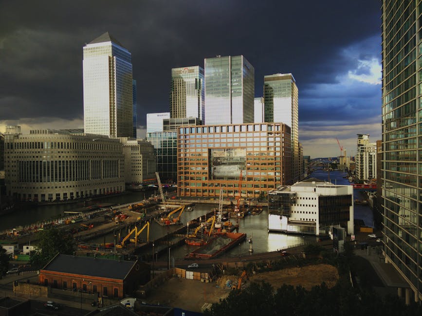 A panoramic view of the Canary Wharf skyline during daytime with modern high-rise office buildings and skyscrapers in the background under a partly cloudy sky. In the foreground, a dock area features