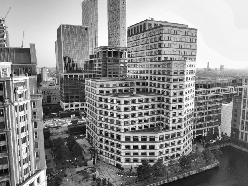 A cityscape at dusk featuring a row of modern high-rise buildings with glass facades reflecting the evening sky, situated along a waterway with calm water and a promenade lined with potted plants and