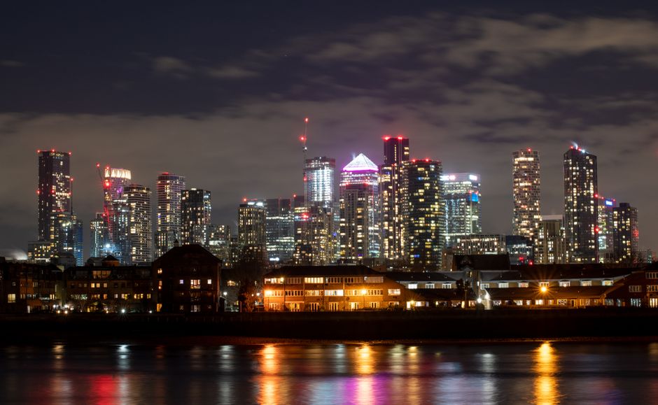 Nighttime view of a modern London city skyline with high-rise office buildings and corporate towers illuminated with multicoloured lights, including red, white, and purple hues, reflecting on the rive