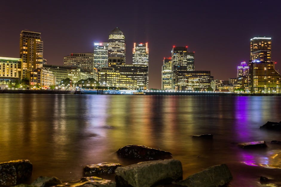A wide-angle view of a canal in Canary Wharf with modern skyscrapers on either side, including glass and steel office buildings and residential apartments. The water reflects the buildings and the par
