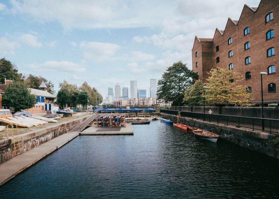A wide outdoor pavement running alongside the River Thames in West India Quay, with a row of mature trees on the right casting dappled shadows on the ground. On the right side of the path, there are r
