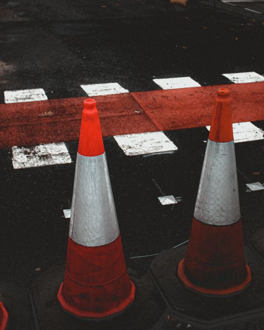 Two orange and white traffic cones placed on a paved street, with a red-marked pedestrian crossing and a red section of road behind them. The cones are situated on the sidewalk close to the curb, near