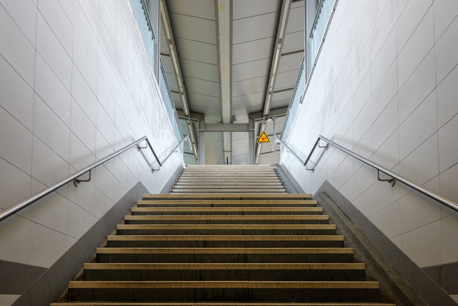 A wide outdoor staircase with multiple steps leading up to a covered walkway at a train or tube station in E14, featuring black and yellow anti-slip safety strips on each step, metal handrails on both